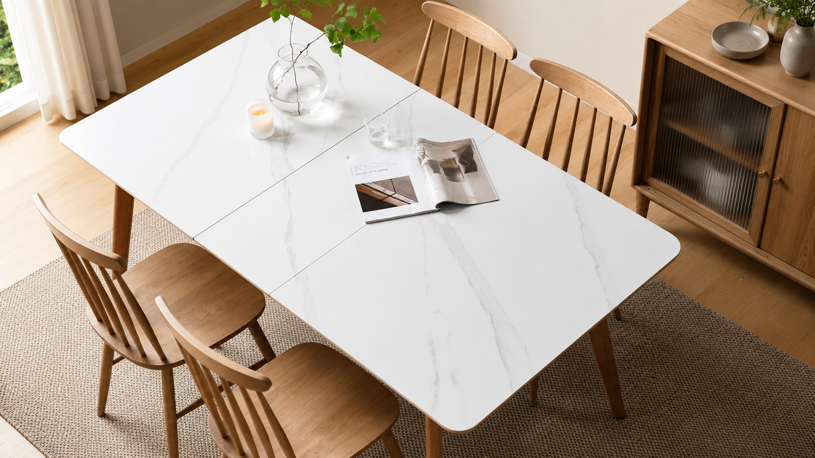 Top-down view of a white marble-top dining table with oak chairs in a bright, modern dining room, styled with a glass vase, candle, water glass, and open magazine.