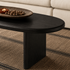 Angled view of a black ash wood oval coffee table with a long rounded tabletop and two solid panel legs.