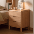 Kusel Natural Solid Oak Bedside Table with three drawers, raised edge top and warm oak grain beside a matching wooden bed.