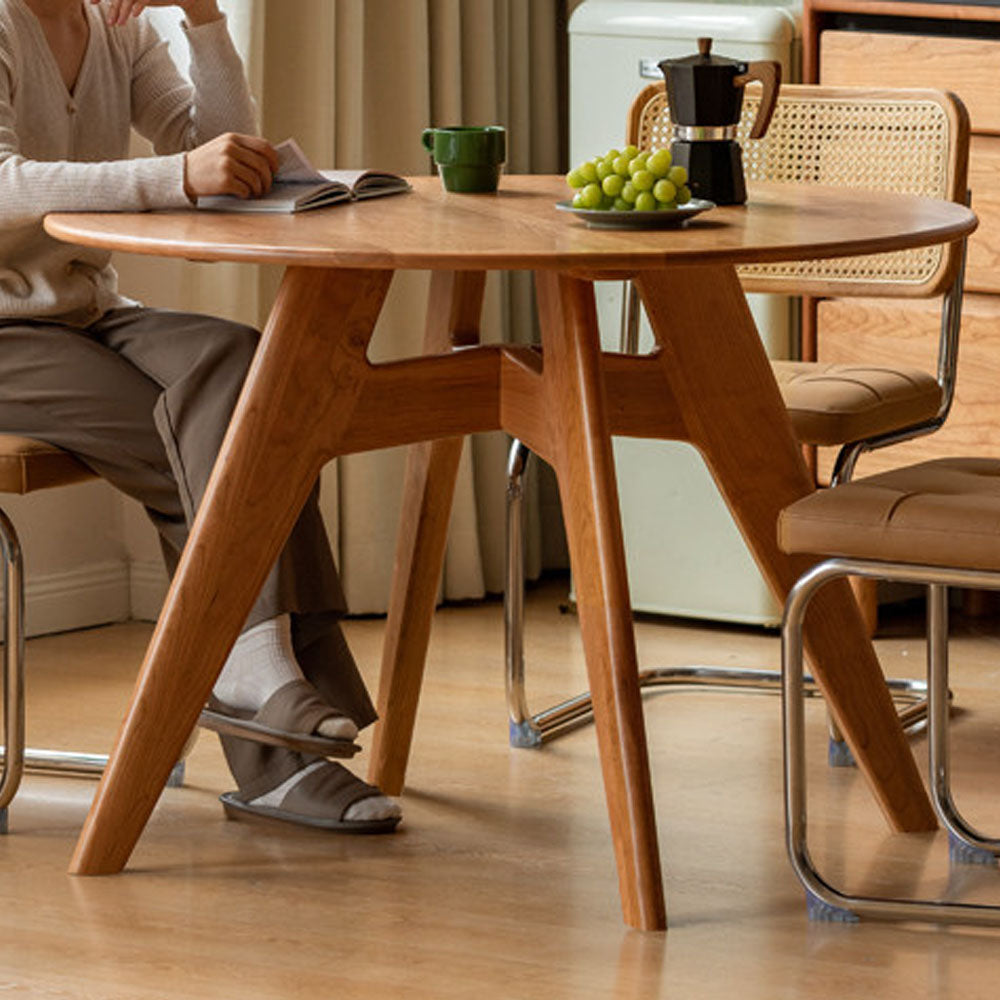 Natural solid cherry small round dining table in a warm timber finish, shown in a compact dining space with dining chairs and kitchen backdrop