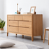 A light oak wood six-drawer dresser styled in a minimalist room with a white wall, gray floor, a potted plant, and a small stool.