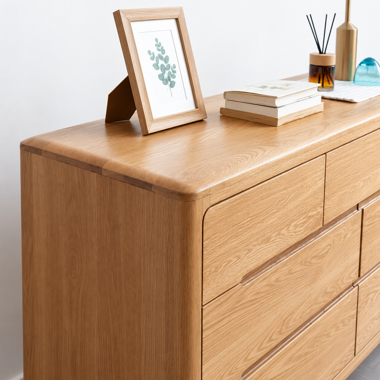 A light oak wood dresser with six drawers, styled with a picture frame, books, and a reed diffuser on top against a plain white wall.