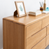 A light oak wood dresser with six drawers, styled with a picture frame, books, and a reed diffuser on top against a plain white wall.