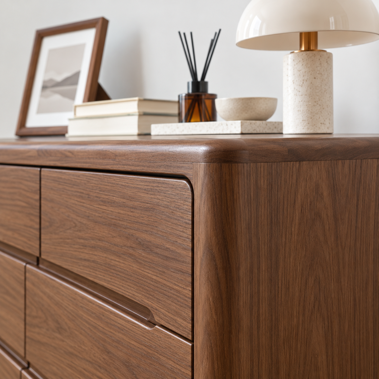 A close-up of a dark walnut wood dresser with six drawers, styled with books, a framed print, a reed diffuser, a small bowl, and a table lamp on top.
