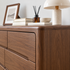A close-up of a dark walnut wood dresser with six drawers, styled with books, a framed print, a reed diffuser, a small bowl, and a table lamp on top.
