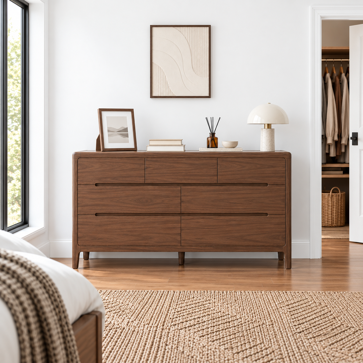 A six-drawer wooden dresser with a warm walnut finish stands in a bright, modern bedroom. The dresser is styled with a small framed picture, books, a reed diffuser, and a white table lamp.