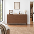 A six-drawer wooden dresser with a warm walnut finish stands in a bright, modern bedroom. The dresser is styled with a small framed picture, books, a reed diffuser, and a white table lamp.