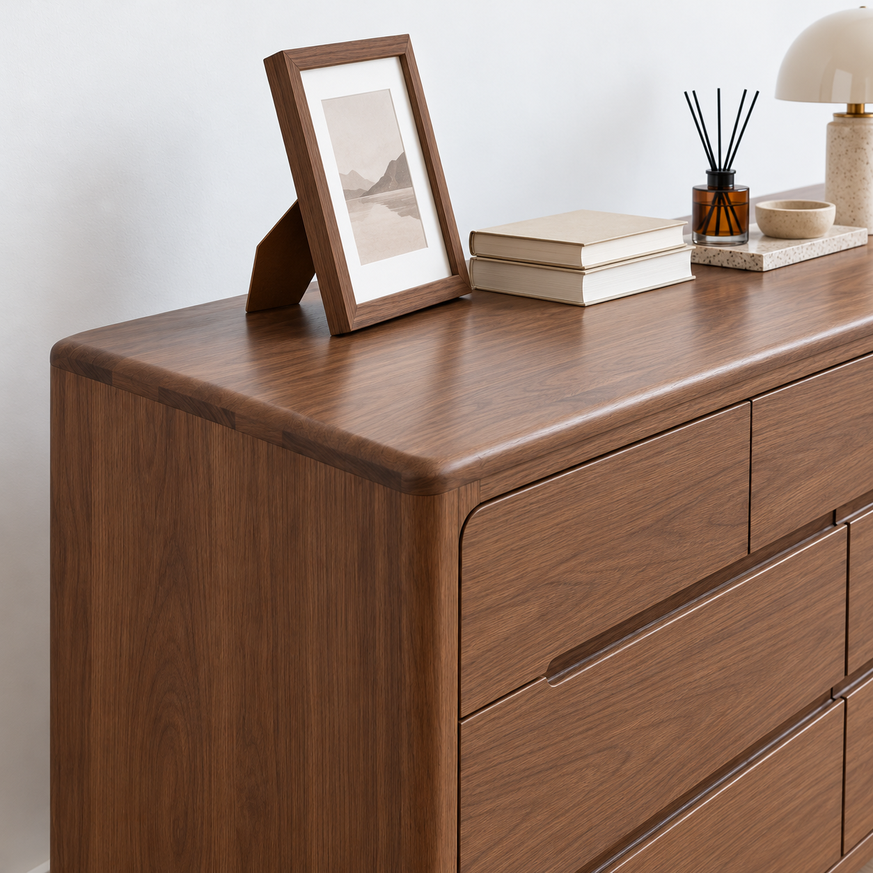 A medium brown wooden dresser with multiple drawers, styled with a picture frame, books, a reed diffuser, and a lamp against a white wall.