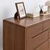 A medium brown wooden dresser with multiple drawers, styled with a picture frame, books, a reed diffuser, and a lamp against a white wall.