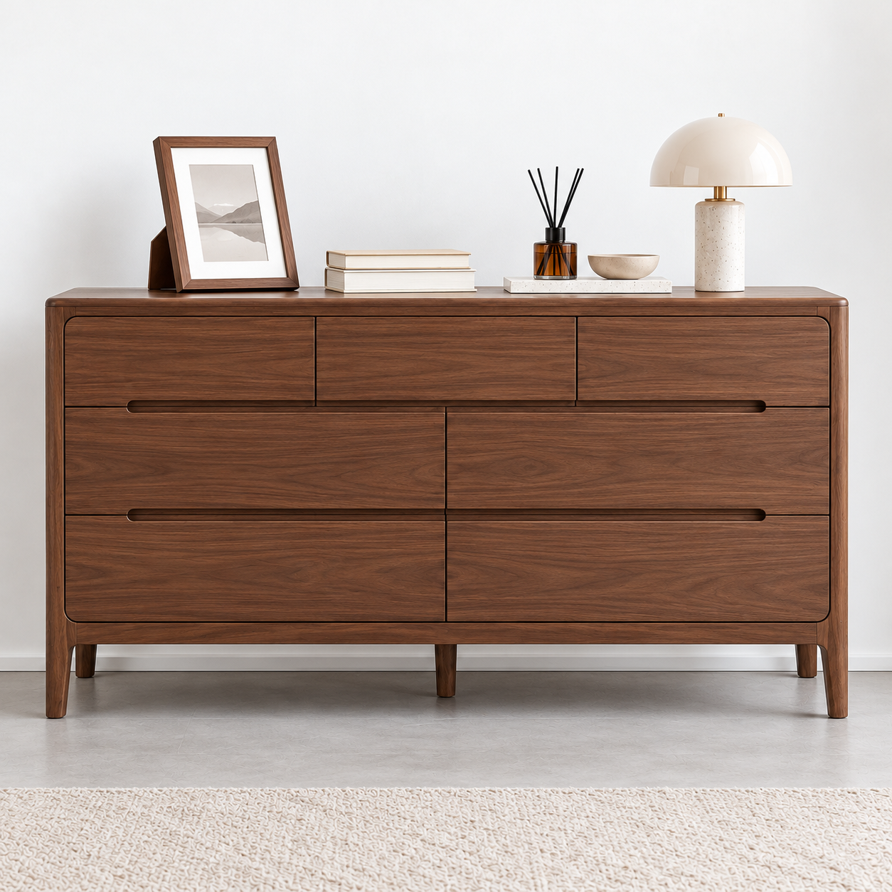A mid-century modern wooden dresser with six drawers, styled with a picture frame, books, a reed diffuser, a small bowl, and a table lamp on top, set against a minimalist white wall and light gray floor.